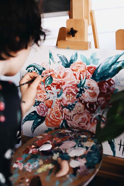 A photo of a lady in a red and floral dress painting while also holding a paint brush and a palette