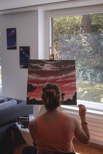 A photo of a woman in a pink tank top evaluating a painted canvas close to a window